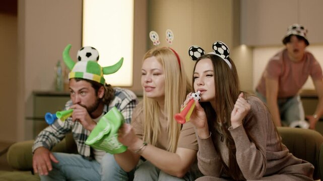 Group of friends celebrating a sporting event at home, wearing festive hats and blowing horns while cheering enthusiastically on a couch in a modern living room