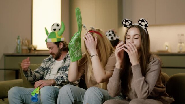 Group of friends wearing festive hats and accessories react to a thrilling moment during a sports event while seated on a couch in a modern living room setting