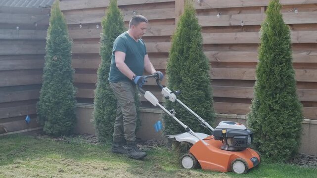 A man uses a professional gasoline scarifier to remove moss and thatch from a green lawn in a private house backyard.