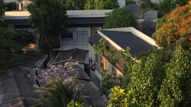 An inviting aerial shot captures a charming resort amidst a canopy of green and flowering trees. Traditional thatched roofs blend with modern architecture under a warm, golden light.