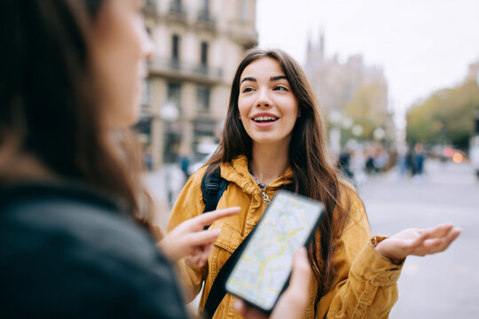 Gen z female traveler asking friend for directions while holding smartphone map, medium close-up street travel moment, lively city background, natural daylight