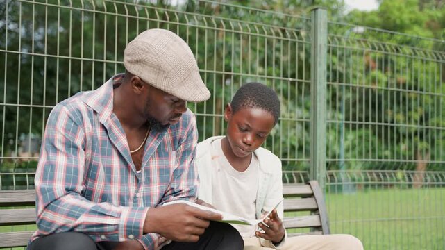 Black father and son drawing on sketchbook while seated on park bench, father points at page and boy smiles, patterned shirt and knit cap, green trees and wire fence backdrop, lighthearted creative