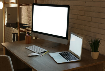 Fototapeta premium Modern programmer's workplace with computer monitor, laptop and shelving unit in office at night, closeup