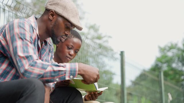 Black father and son writing together on notebook while seated on park bench, father demonstrates handwriting stroke and boy concentrates, knit cap and plaid shirt, wire fence and leafy trees