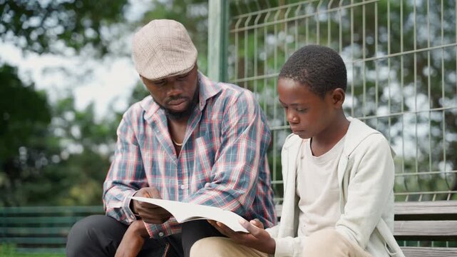 Park bench father and son reading, man in plaid shirt and cap and boy flip through open book together, calm instructional moment, trees and fence backdrop, shared smiles, educational bonding
