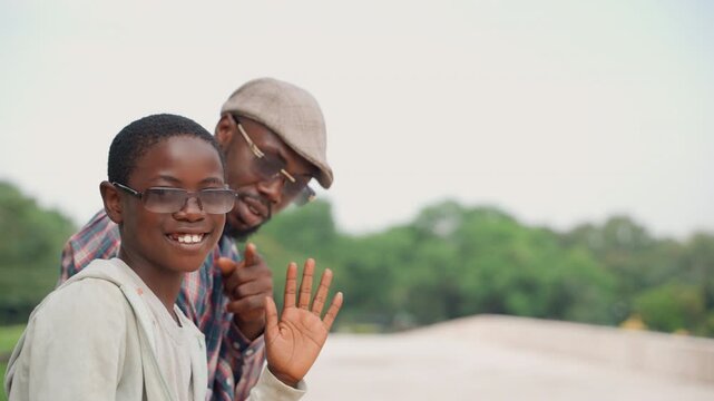 Sunlit park black father and son sharing playful wave and bright smiles, son wearing glasses and cap while father peeks over shoulder, candid joyful moment with greenery backdrop and breezy atmosphere