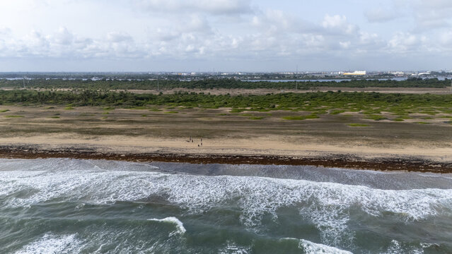 Aerial view of the coastline featuring crashing ocean waves, a sandy beach with a few people, and lush green vegetation under a cloudy sky in Badagry, Lagos, Nigeria.