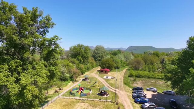 Aerial drone forward flight over a riverside park near Nis Serbia with a colorful playground outdoor basketball court parked cars lush green trees and distant mountain ranges on a sunny spring day