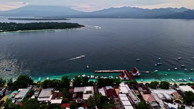 Sweeping drone aerial of a large coastal city with dense urban development, mosque visible, fronted by a long dark sandy beach and backed by dramatic mountain ranges. Lombok, Indonesia, Kota Mataram.