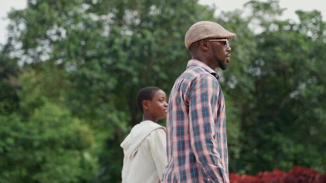 Father and son walking in park, casual plaidshirt dad beside young boy in hoodie, sideprofile shots, leafy trees and red shrubs in background, overcast sky, slow intimate bonding moment, gentle pace,