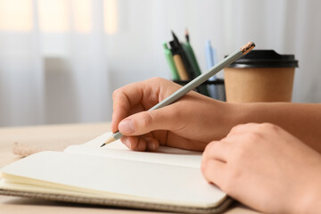 Fototapeta premium Woman with pencils holder and cup of coffee writing something in notebook at workplace, closeup