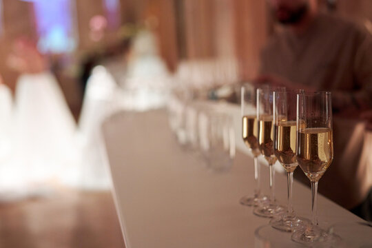 Champagne Glasses in a Row at Luxury Wedding Reception. Close-up of sparkling champagne flutes lined up on a white bar counter during a gala event or wedding. Blurred festive background