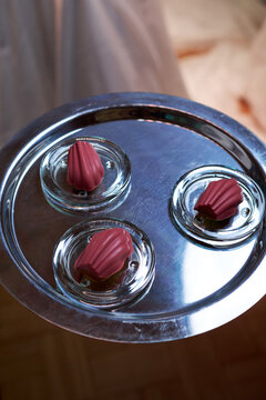 High-angle shot of three pink chocolate-glazed madeleines served on glass saucers on a silver tray. Elegant gourmet dessert catering for a luxury banquet, wedding, or corporate event
