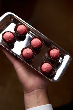 Close-up of a waiter hand holding a silver tray with pink chocolate truffles in brown paper cups. Elegant dessert catering for a luxury banquet, wedding reception, or event
