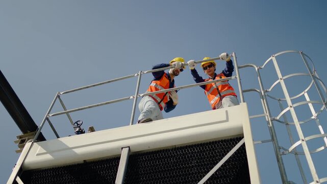 Two professional HVAC technicians in safety gear and hard hats inspect an industrial cooling tower on a rooftop under a blue sky, communicating via walkie-talkie for maintenance and engineering servic
