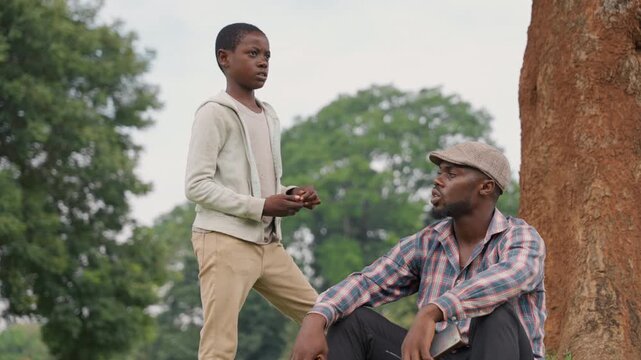Black father and son under tree sharing quiet conversation, father seated on grass, son standing with hands clasped, oak backdrop, soft overcast light, intimate mentorship, rural park setting, calm