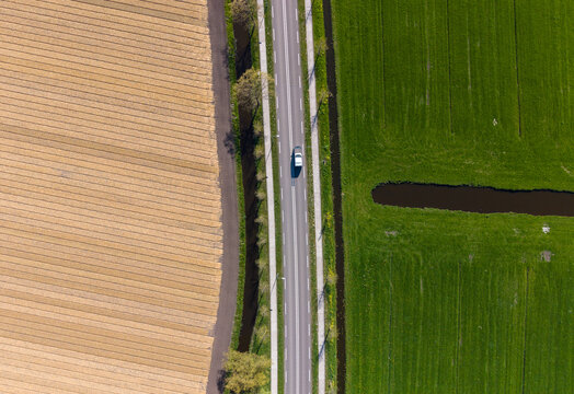 Aerial view of a white car driving on a straight road between contrasting brown and green agricultural fields in Heiloo, North Holland, Netherlands.