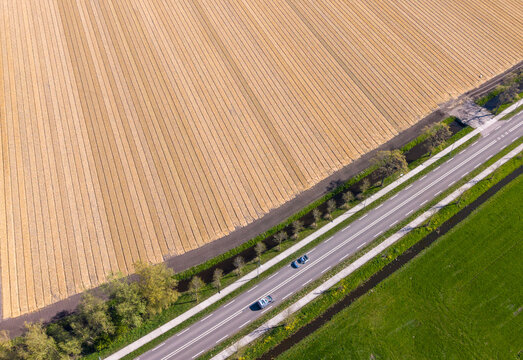 Aerial view of a paved road with cars passing between a vast agricultural field with parallel rows and a lush green meadow in Heiloo, North Holland, Netherlands.