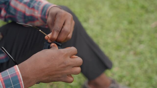 Outdoor man polishing lens carefully. Relaxed individual in plaid shirt cleaning spectacles outdoors carefully. Casual man wearing plaid attire gently polishing eyeglasses on bench outdoors