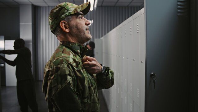 Male Soldier With Tattooed Arms Puts on Camouflage OCP Uniform Jacket and Cap by Locker, Getting Ready For Tactical Mission While Colleagues Interact in Background. Concept Military Base Preparation.