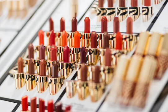 Display of various lipstick shades arranged in rows on a cosmetic shelf, showcasing a range of colors from reds to nudes with gold packaging in a beauty store setting