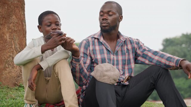 Black father and son sitting under tree sharing quiet conversation, father acting as mentor while son listens thoughtfully, park grass and warm afternoon, candid emotional portrait of guidance