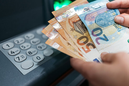 Hands holding various euro banknotes after a transaction at an ATM, with a keypad and green light visible in the background