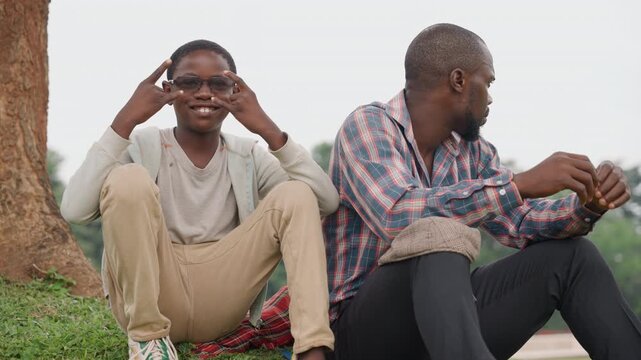 Black father and son sitting under tree, casual mentor and teen share quiet conversation on ground, father checks phone while son gestures with sunglasses, relaxed park atmosphere, intergenerational