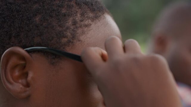 Smiling black son wearing sunglasses outdoors, proud youth beams after trying on shades while father rests nearby, sunlight highlights cheek and confident grin, playful moment of self discovery