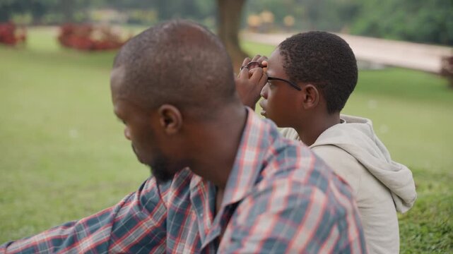 Park black father and son sitting side by side on grass, father in plaid shirt, son in hoodie, quiet conversation and shared silence, generational connection, casual afternoon, candid lifestyle moment