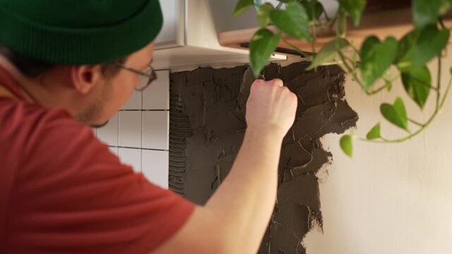Young man in a beanie applying tile adhesive to a kitchen wall with a putty knife for a backsplash installation, showcasing the process of home improvement and do-it-yourself renovation work