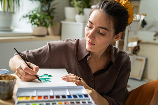 Pensive woman artist enjoying new watercolor paints, drawing sketches in sketchbook at home studio, focused positive female painter sitting relaxed on floor, painting in paper book with aquarelle.