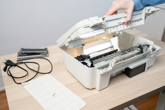 Person performing maintenance on an open white inkjet printer scanner on desk, showing internal components, power cable and removed parts, illustrating repair, troubleshooting and office technology.