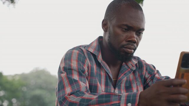Black father staring at phone alone under tree in park, pensive expression, checking messages with focused gaze, patterned shirt, soft bokeh background, quiet tension and introspective mood