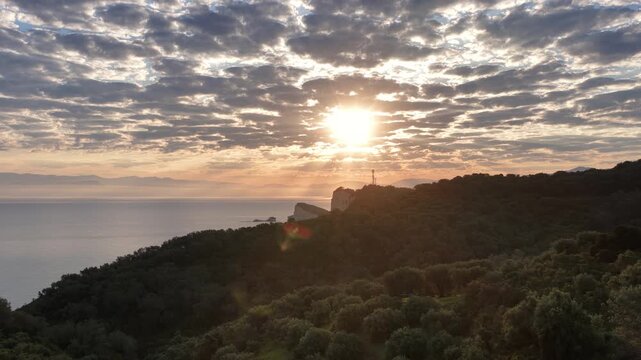 Breathtaking morning sun shining through patterned clouds over Cape Drastis, Corfu Greece. Aerial drone view of dramatic Mediterranean coastline, steep cliffs, and golden sunlight on the sea.