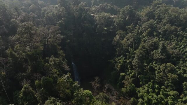 Th&aacute;c Ti&ecirc;n waterfall drifting drone view over rainforest treetops, with the ravine and cascade partially obscured by haze, H&agrave; Giang, Vietnam.