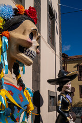 Obraz premium Colourful Catrina decorated with flowers and ribbons. Skeletal figure wears festive attire. Second figure stands in dark formal garb. Bright blue sky contrasts with aged buildings