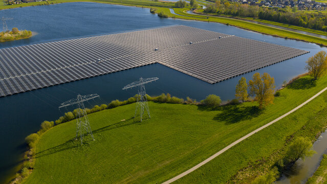 Aerial view of the Sekdoornse Plas floating solar farm with rows of photovoltaic panels on the water and power pylons on the green shore in Zwolle, Overijssel, Netherlands.