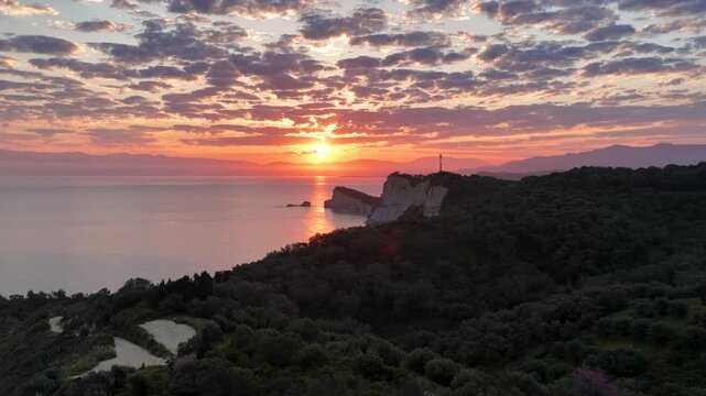 Aerial view of colorful sunrise over Cape Drastis in Corfu Greece. Dramatic pink clouds, steep rocky coastal cliffs, and lush green hills meeting the calm Mediterranean Ionian sea.