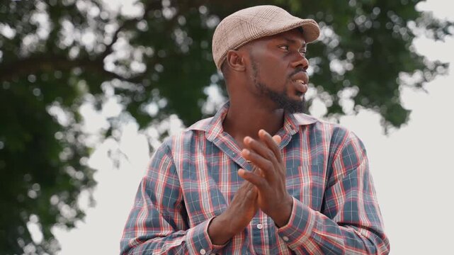 Outdoor man speaks with animated gestures, cap and patterned shirt under leafy canopy. Series of dynamic moments turning head, clapping hands, emphasizing points, briefly inspecting a small object,