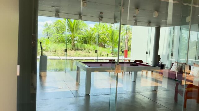 Modern luxury game room with professional pool table and panoramic glass walls overlooking tropical garden.