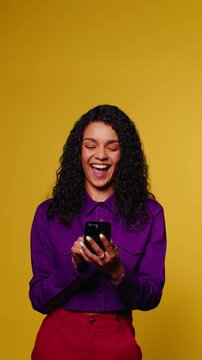 A young woman with curly hair scrolls and laughs at something on her phone against a vibrant yellow background in a vertical medium shot.