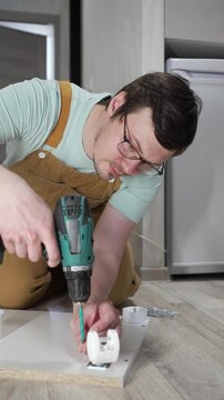 Concentrated male worker in overalls using a cordless screwdriver to assemble flat-pack furniture, meticulously fastening components for a new cabinet during a home renovation