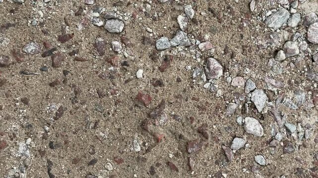 Close-up worn concrete road surface showing gritty pebbles and rubble, weathered asphalt fragments, fine sand and loose stones.