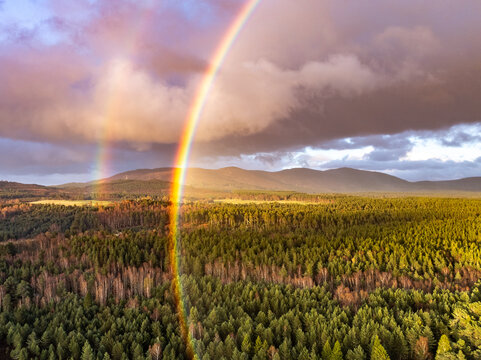 Aerial view of Cairngorms National Park featuring a vibrant double rainbow over a dense evergreen forest and rolling mountains under a dramatic sky in Scotland, United Kingdom.
