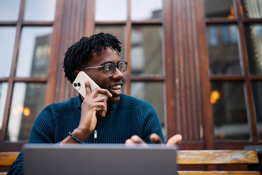 Happy African American freelancer talking on mobile phone while working laptop in outdoor cafe.