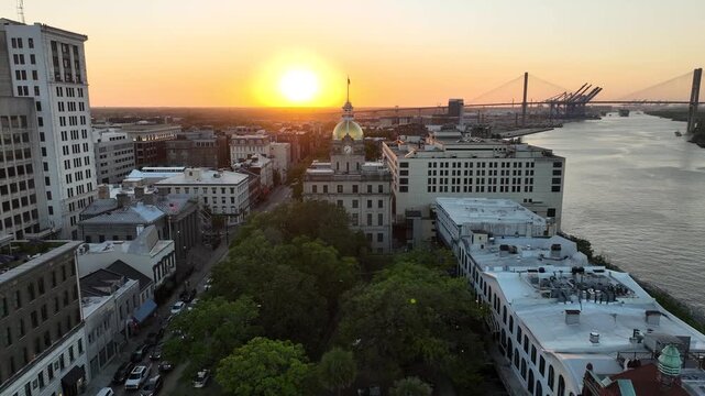 Breathtaking sunset drone shot of Savannah, Georgia, capturing the architectural beauty of City Hall's domed clock tower nestled among historic buildings, with the Savannah River and cargo port stretc