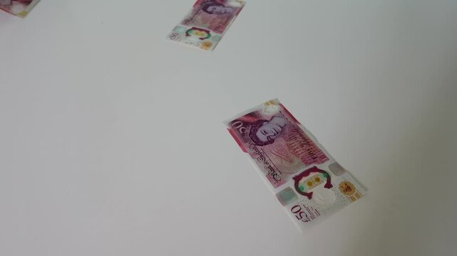 Multiple British polymer &pound;50 notes cascade and fall at random onto a table surface in 120fps slow motion, capturing every twist and flutter of each note in detail.