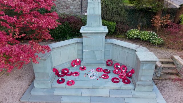 A reverse rising drone shot pulls back from a war memorial adorned with poppy wreaths in Windermere memorial gardens, revealing Cumbrian slate cottages, spring blossom and the Windermere to Bowness Rd