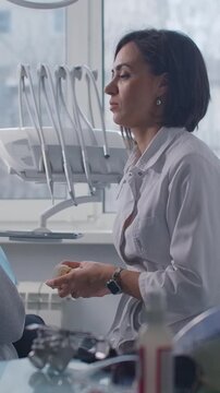 Female dentist consulting with patient in modern dental clinic, explaining treatment using tooth model while reviewing digital x-ray results on computer monitor